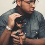 A man in casual attire sits indoors with a black Chihuahua, showcasing a warm interaction.